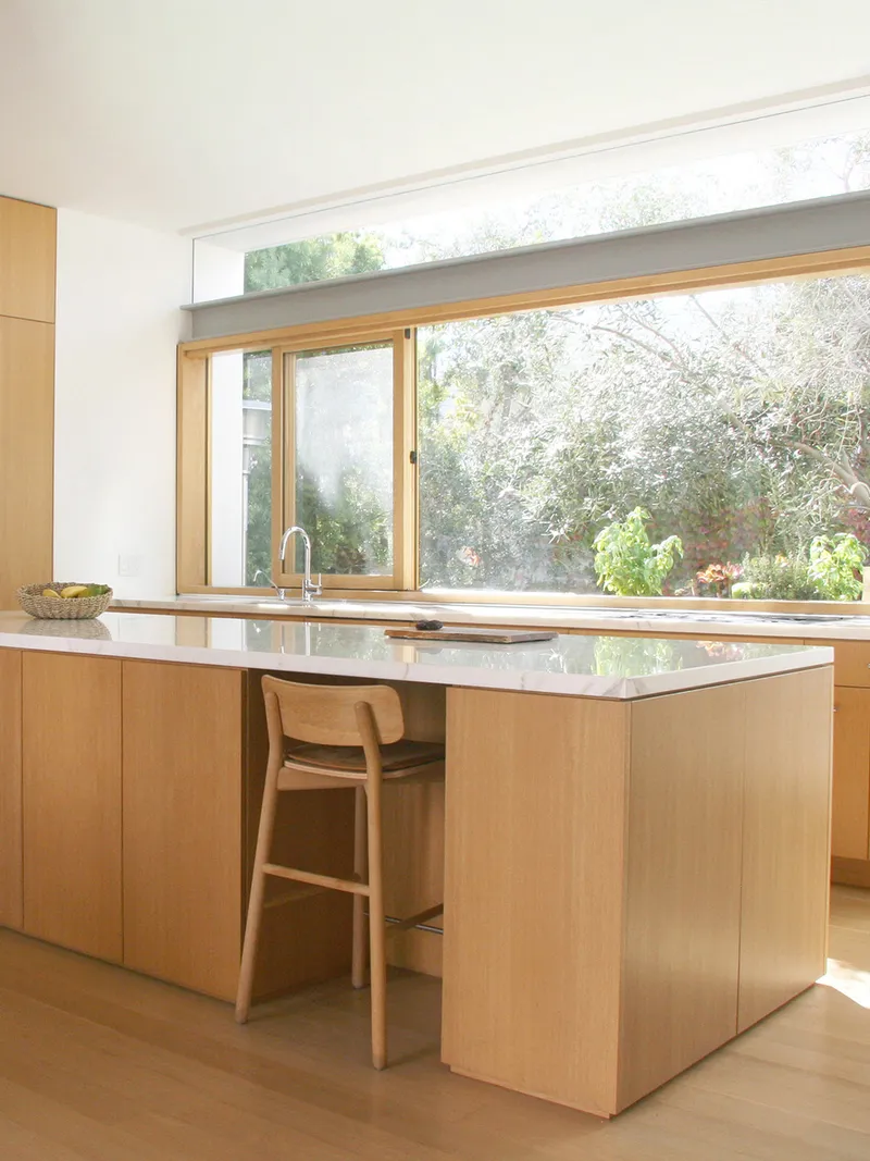 Kitchen island with white stone countertop facing timber-framed windows overlooking a garden