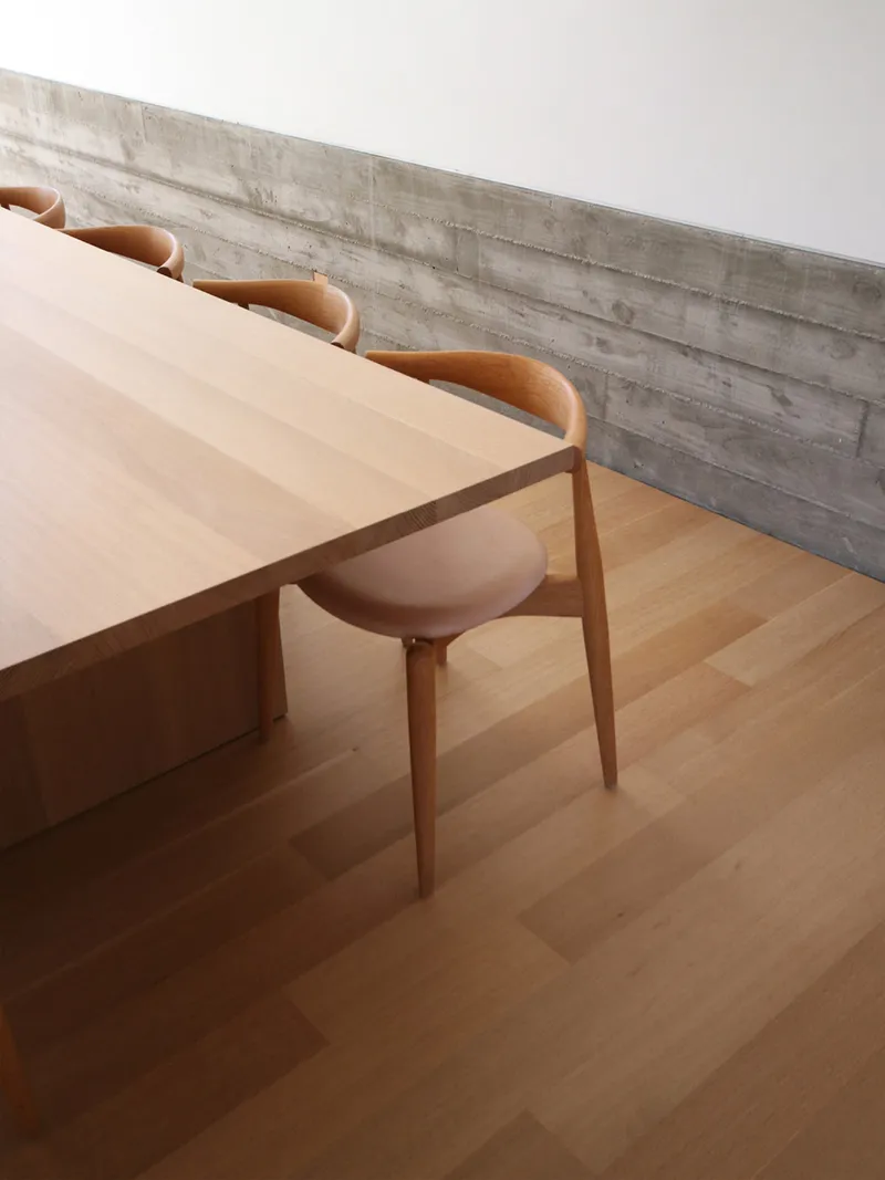 Light timber dining table and chairs against a board-formed concrete wall in soft natural light