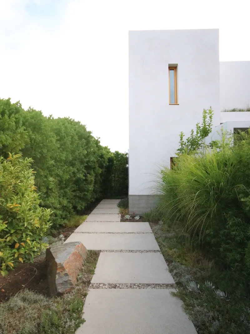 Concrete paver path flanked by grasses and plantings leading to a white stucco tower volume