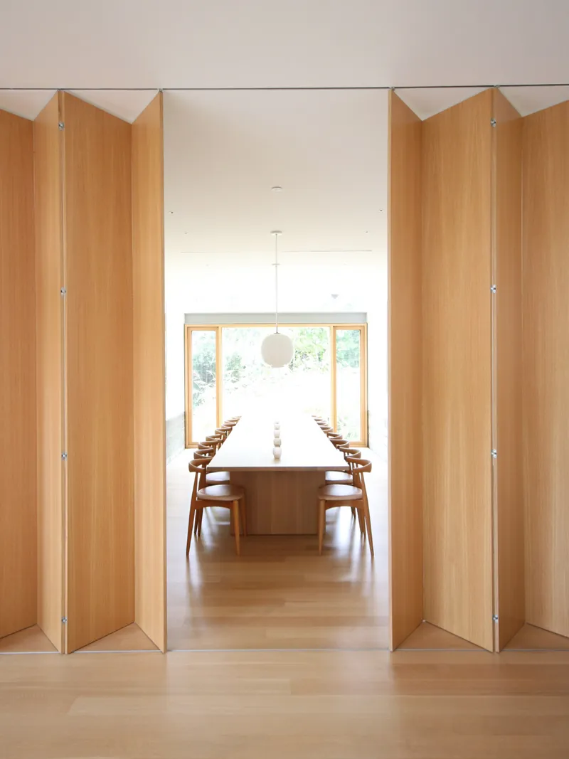 View through open angled timber doors toward a dining room with long table under pendant light