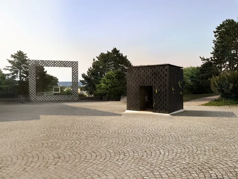 Two basketweave brick pavilions on a cobblestone plaza at dusk with trees in the background