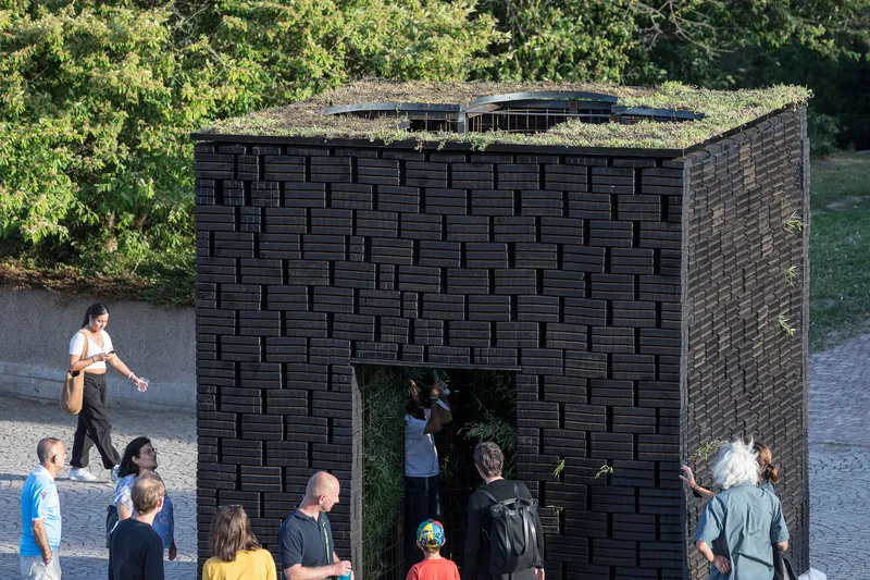 Facade of woven dark brick pavilion with planted roof and visitors gathering near the entrance