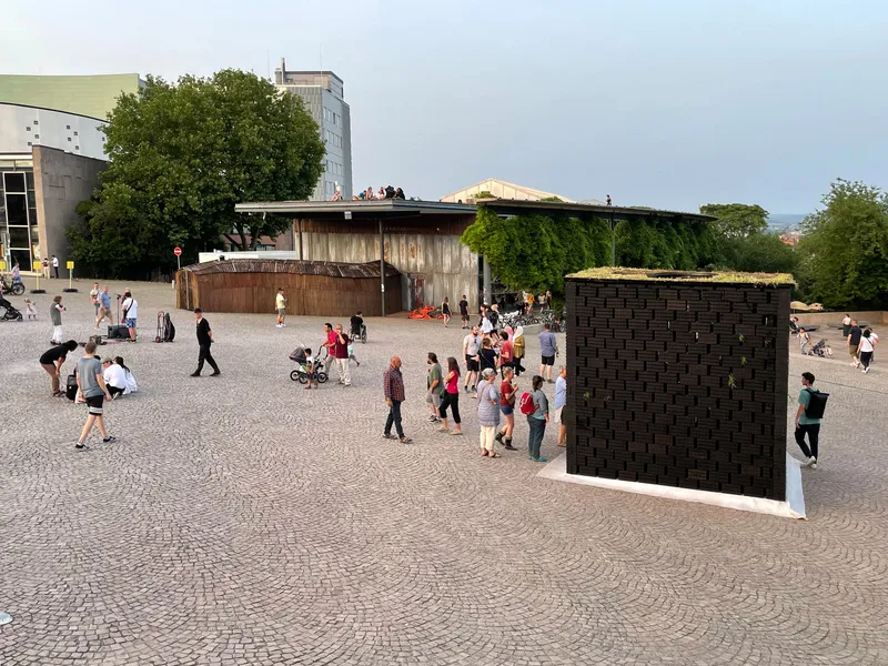 Public plaza with timber-clad pavilion and woven brick volumes amid pedestrians on patterned paving