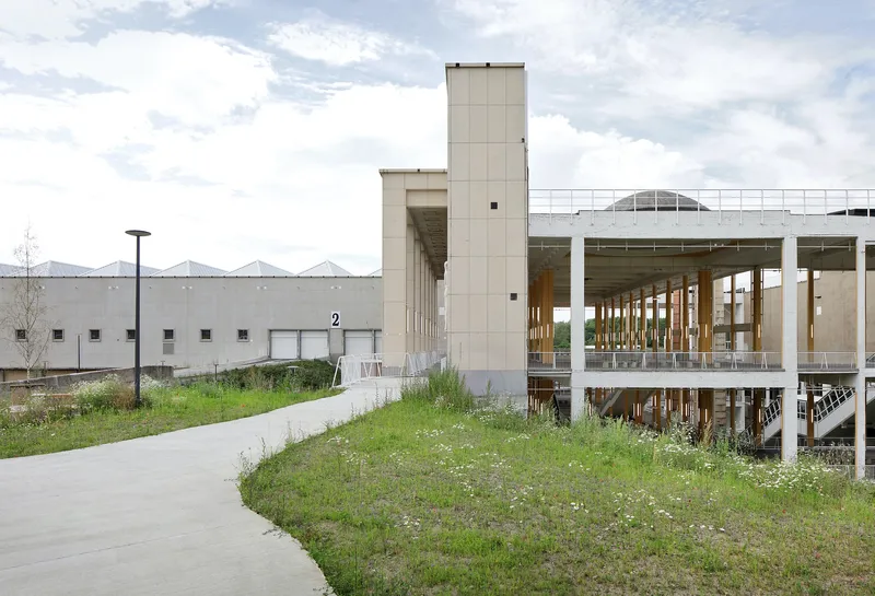 Elevated glass-walled pavilion with exposed timber columns beside a grassy path under cloudy skies