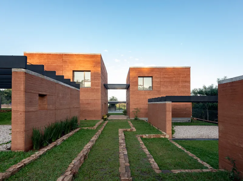 Front courtyard with rammed earth walls, planted grass strips and gravel paths leading to a skybridge