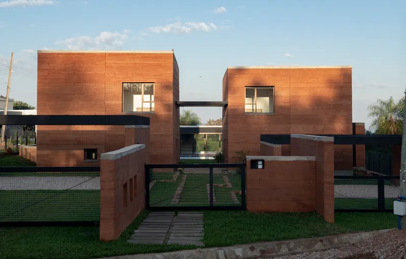 Street view of paired rammed earth volumes connected by a bridge under evening light