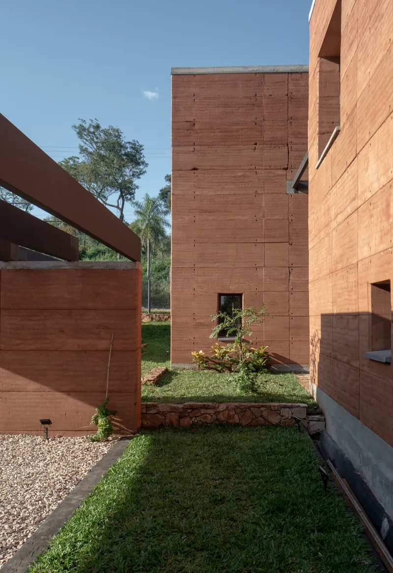 Narrow garden passage between rammed earth volumes with grass strips and young planted trees