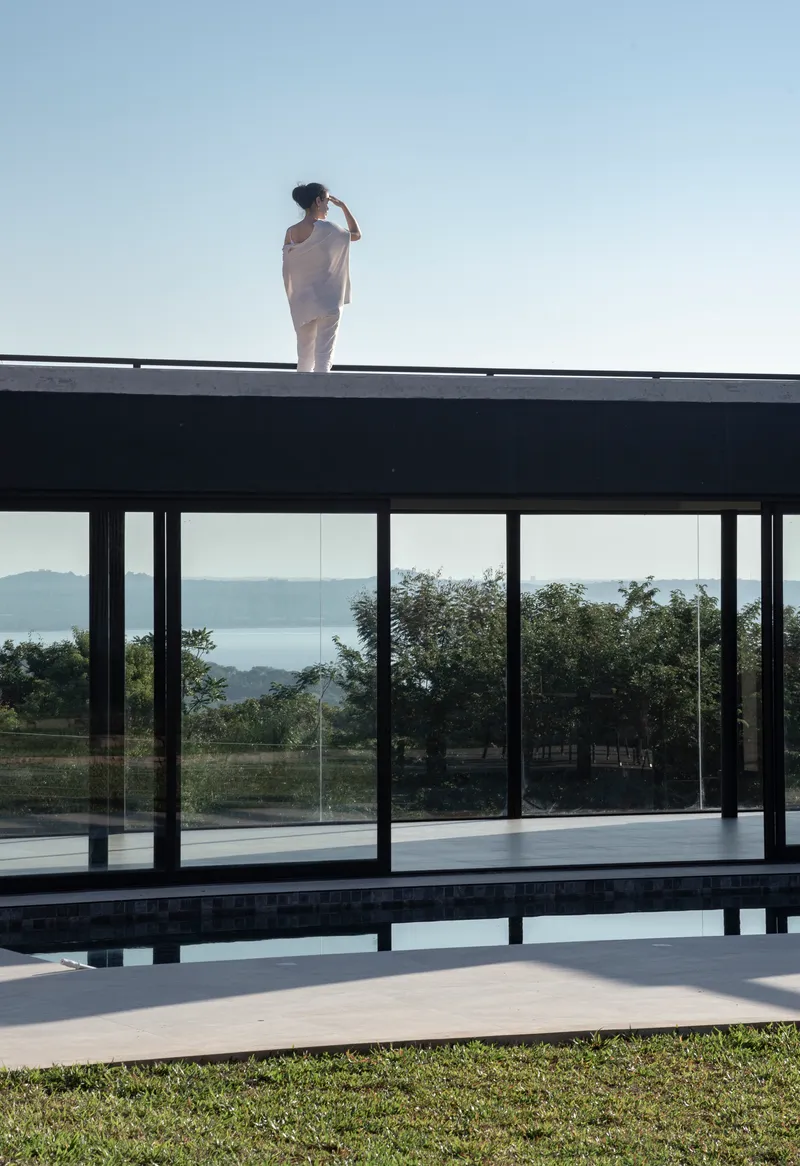 Figure standing on the flat roof above the glass-walled pool pavilion overlooking distant hills