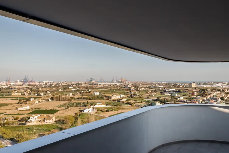 Curved balcony edge beneath dark overhang overlooking rural landscape with industrial skyline in the distance