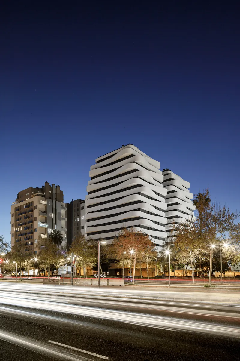 Street view of the stacked balcony facade at twilight with traffic light trails and illuminated trees