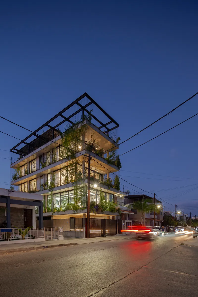 Corner street view at dusk showing planted balconies and rooftop steel frame with passing car lights