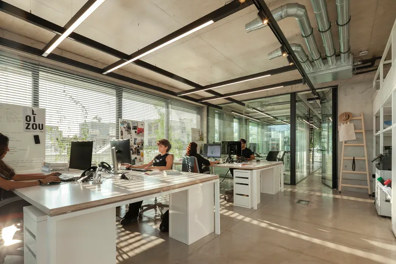 Open office interior with exposed concrete ceiling, steel beams, and workers at white desks