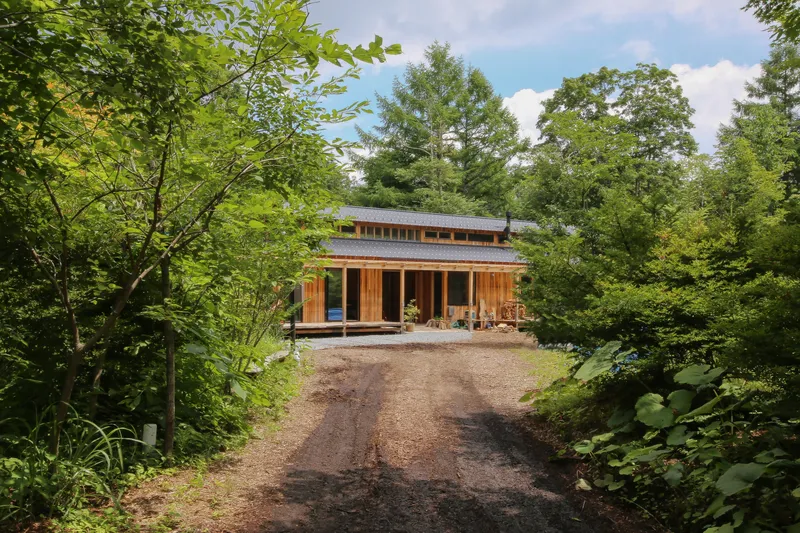 Two-story timber structure with wraparound porch and clerestory band viewed from gravel approach through summer foliage