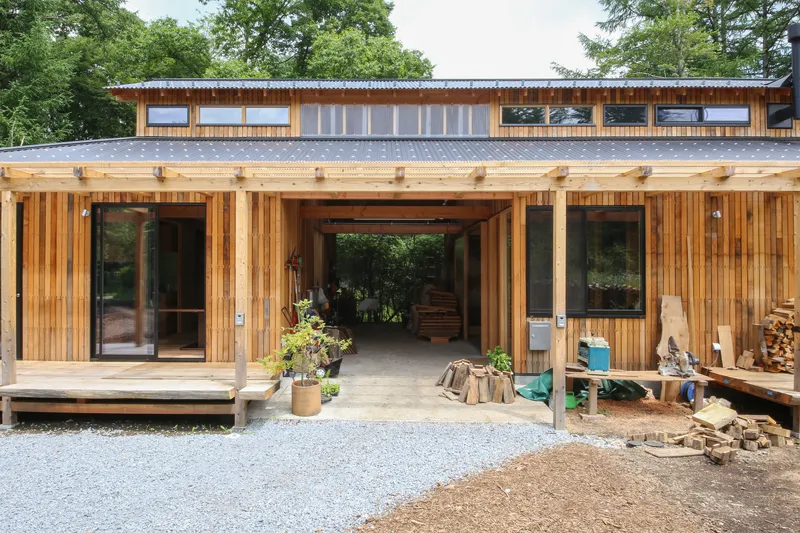 Vertical timber slatted facade with central open passageway and gravel forecourt surrounded by mature trees
