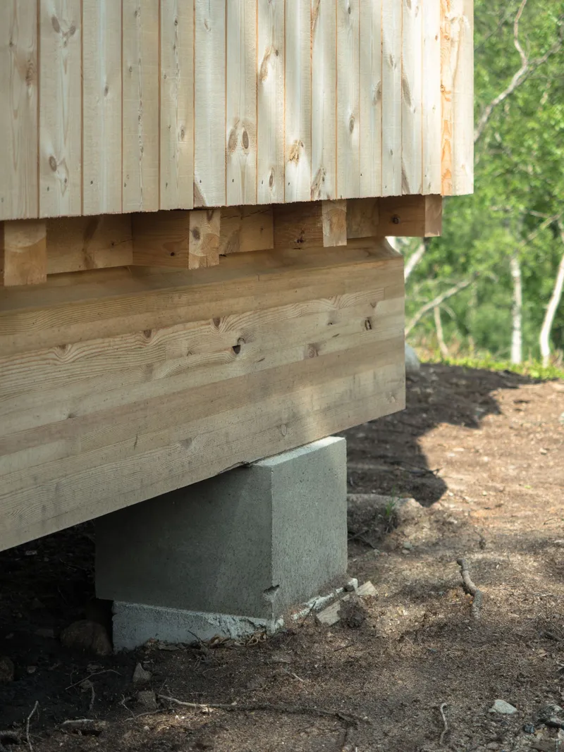 Foundation junction showing horizontal wood cladding supported on concrete plinth with surrounding earth and vegetation