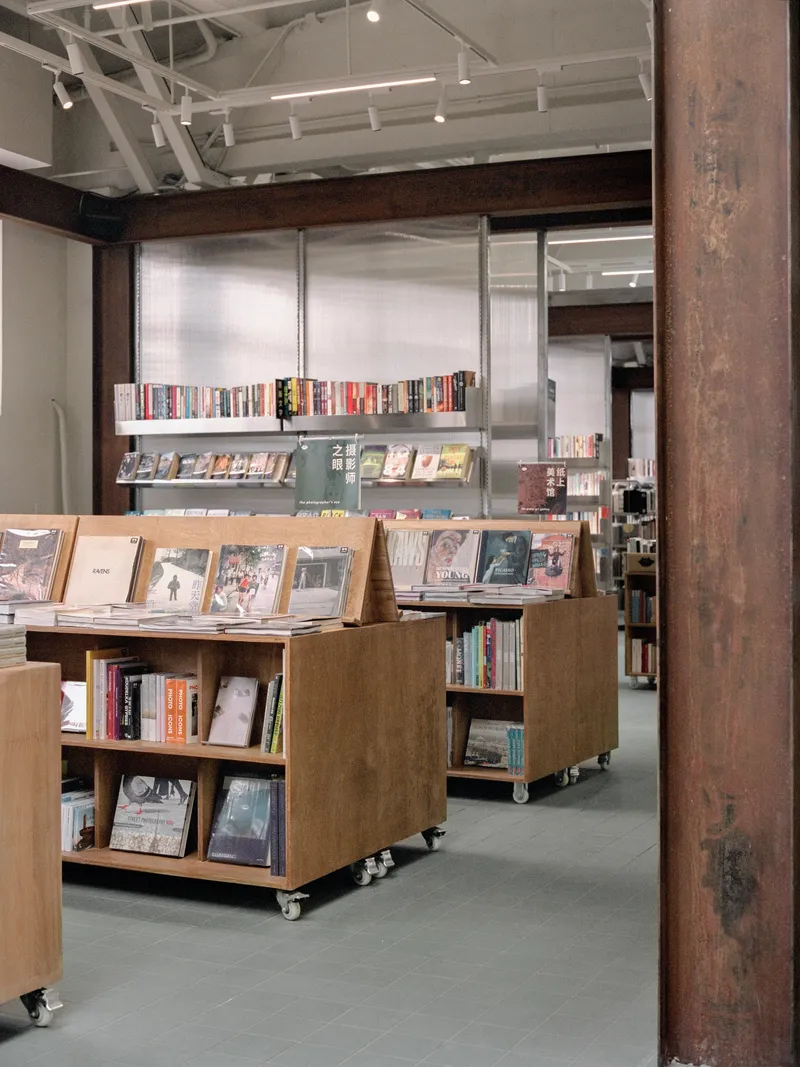 Wheeled plywood display units holding books in front of translucent wall panels and metal shelves beyond timber column