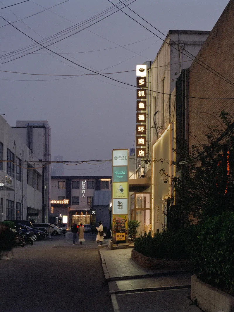 Narrow street view at dusk with illuminated storefronts and overhead power lines
