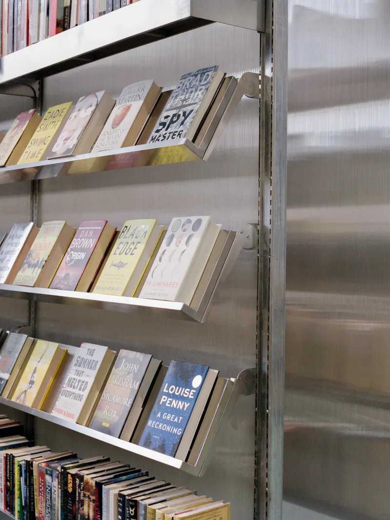 Metal bookshelves with angled display ledges holding books against a corrugated polycarbonate wall