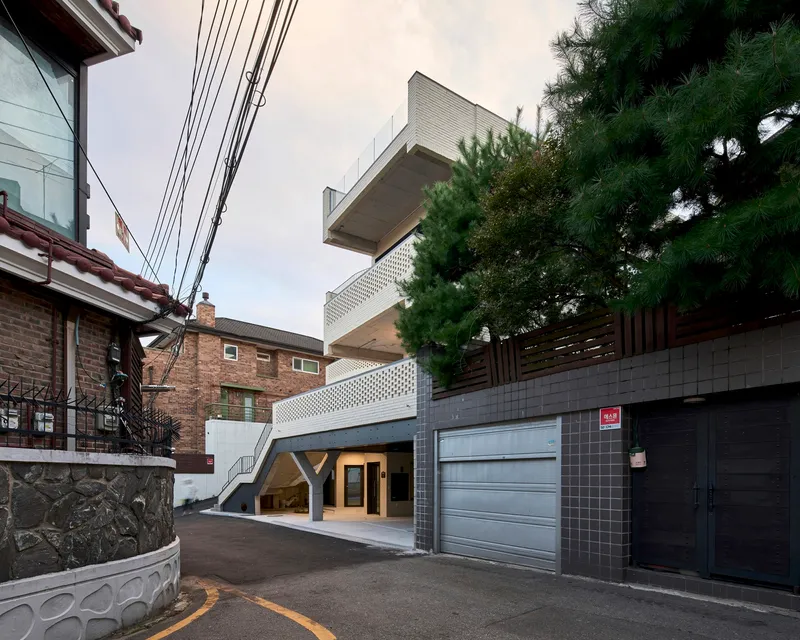 Street view showing stacked volumes with perforated screens and cantilevered terraces beside power lines and pine tree