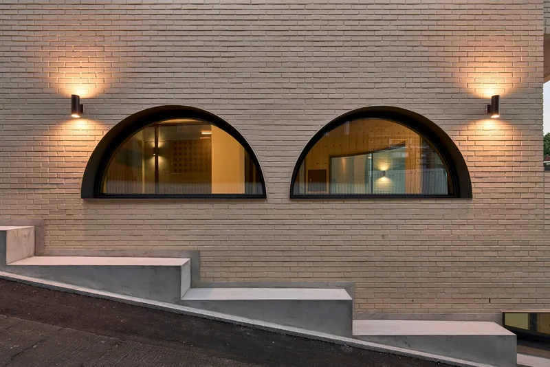 Two arched windows with wall sconces on cream brick facade above stepped concrete terrace at twilight