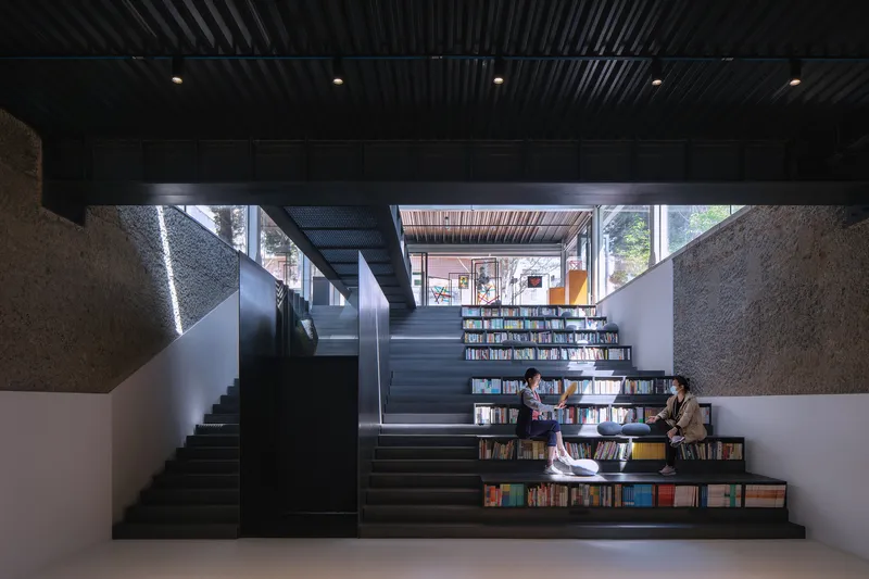 Stepped seating staircase integrated with bookshelves where two readers sit beneath a corrugated metal ceiling