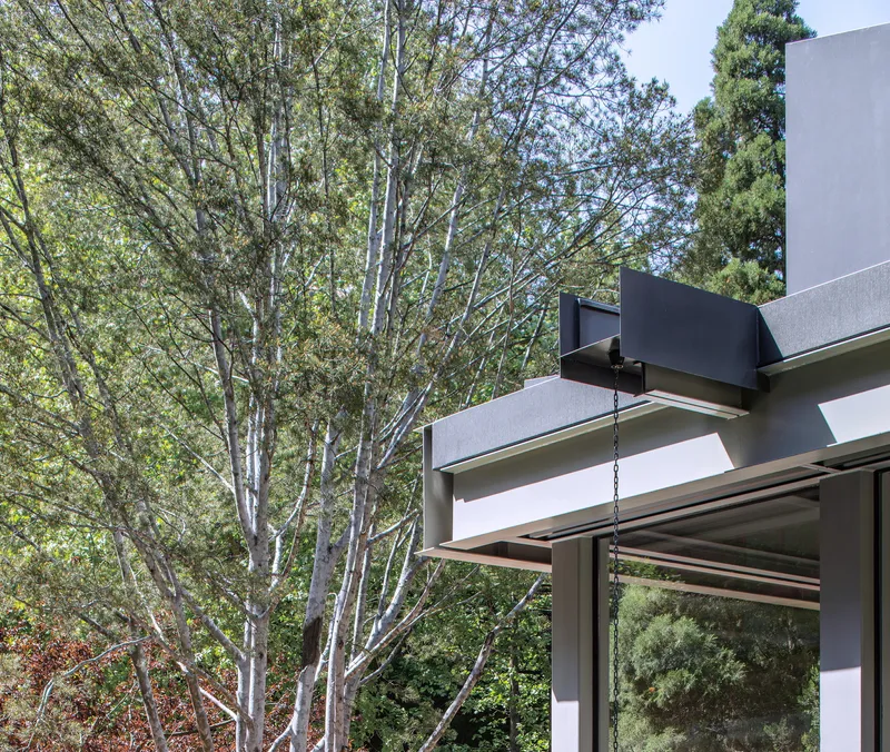 Close-up of the black metal roof connector and white beams with tree branches visible through glass