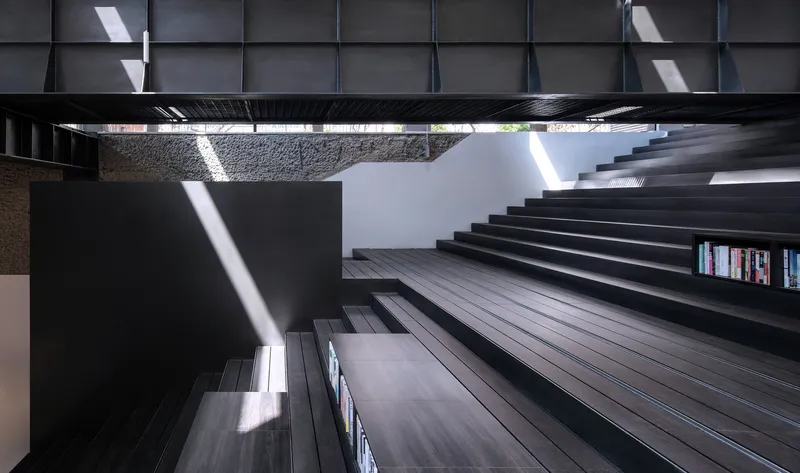 Looking up through the central atrium at black timber stair treads bridging between stone walls with sunlight streaming down