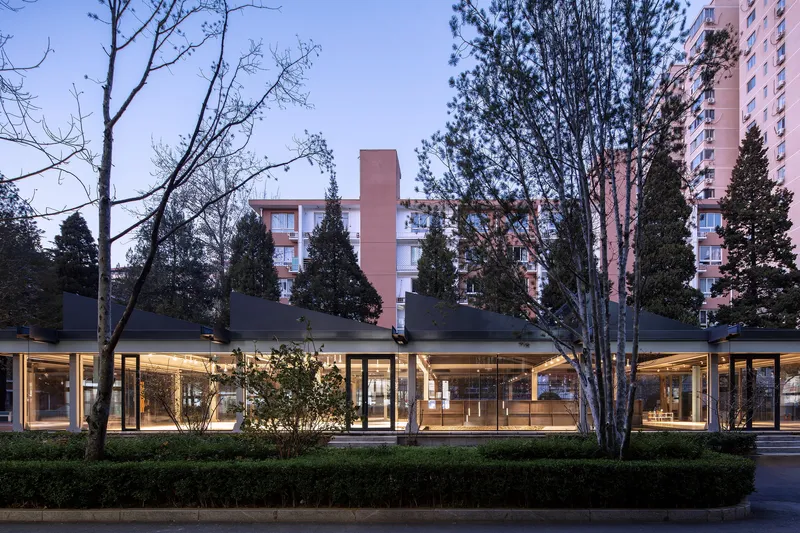 Exterior view of the low glass pavilion with overhanging roof among bare trees and residential towers at dusk