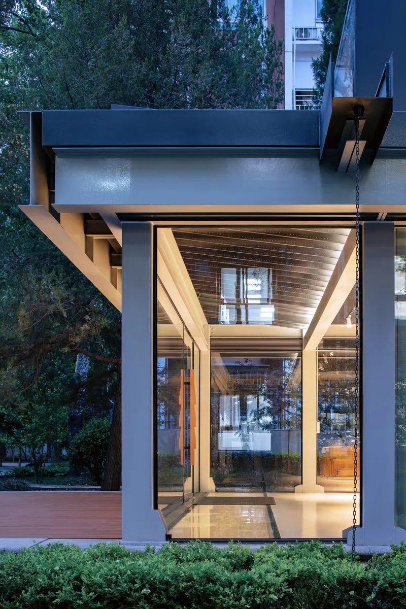Glass pavilion with exposed timber rafters and white columns framed by dense foliage at dusk