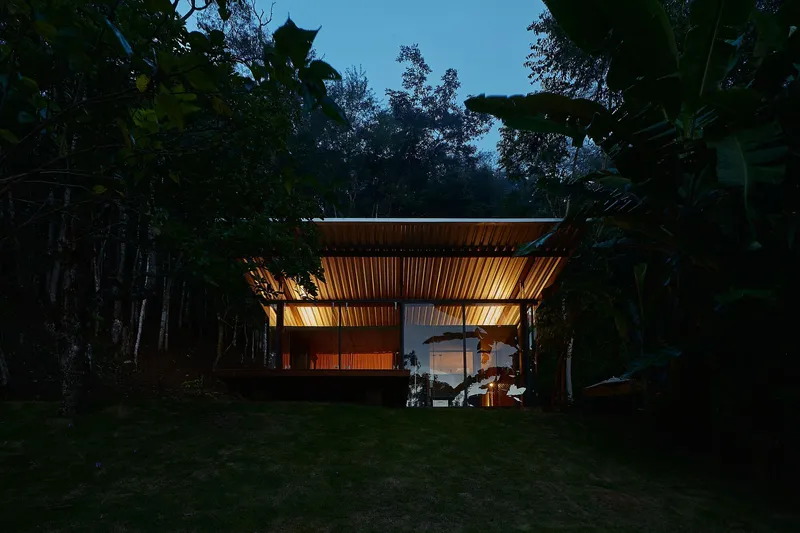 Illuminated pavilion with horizontal timber slat screens under a flat roof at dusk in a forested clearing