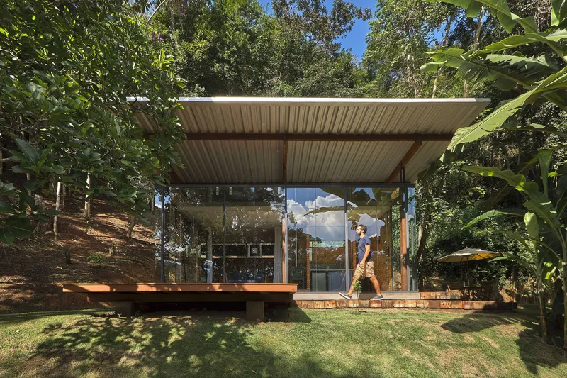 Glass pavilion with corrugated metal roof and timber deck set in a sloped clearing surrounded by trees