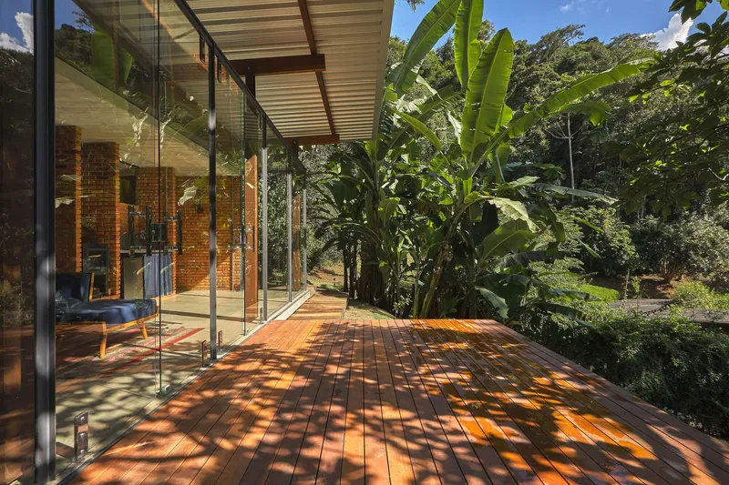Timber deck with dappled sunlight filtering through overhanging trees beside a glazed wall and exposed brick interior