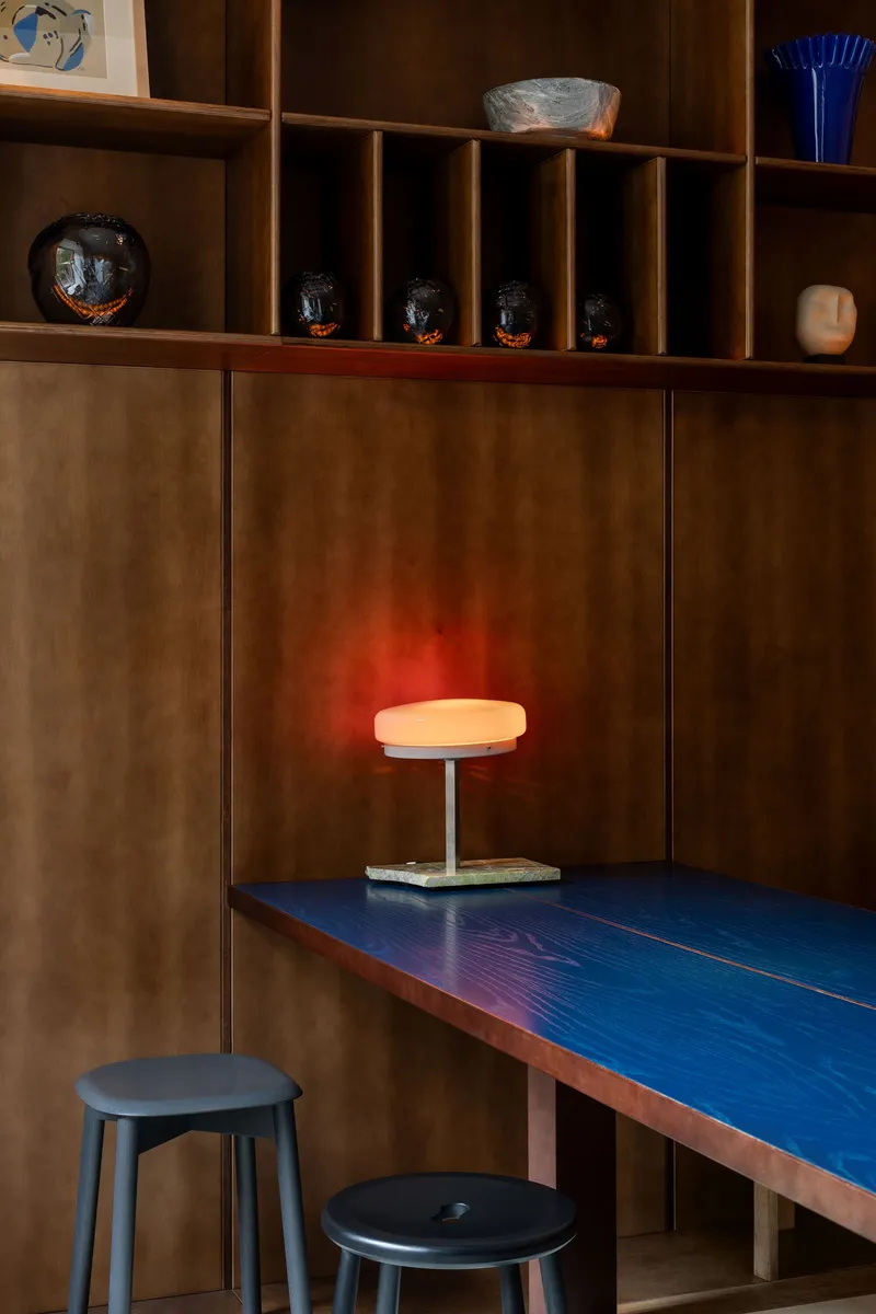 Built-in desk with blue laminate surface, glowing lamp, and timber shelving displaying ceramic objects