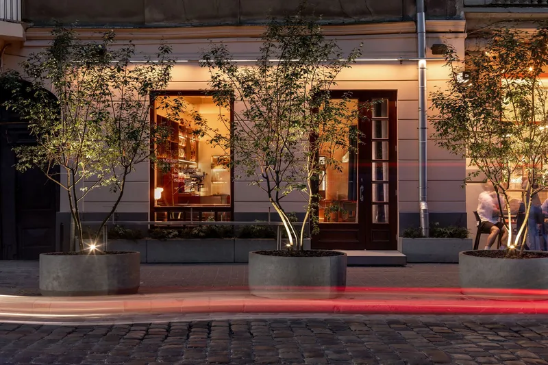 Street facade at dusk with concrete planters, uplighting on young trees and blurred pedestrians passing