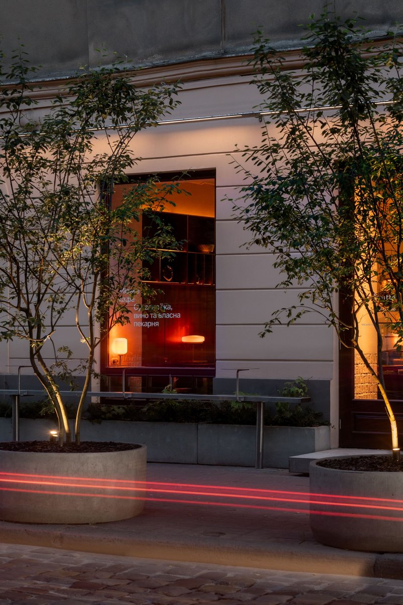 Street entrance with uplighted trees in concrete planters and warm interior glow at dusk