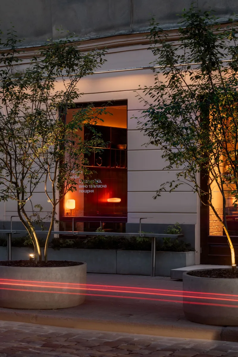 Street entrance with uplighted trees in concrete planters and warm interior glow at dusk