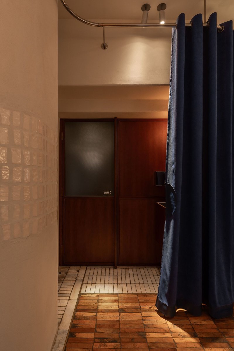 Corridor with curved brass curtain rail and clay tile floor leading to a walnut restroom door