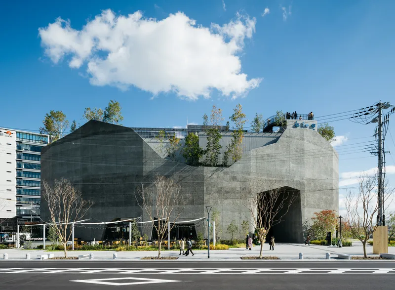 Street view of the faceted concrete volume with rooftop planting and pedestrians crossing below a blue sky