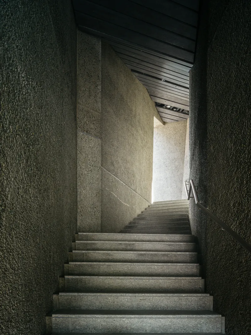 Narrow concrete staircase ascending between textured aggregate walls toward a bright opening above