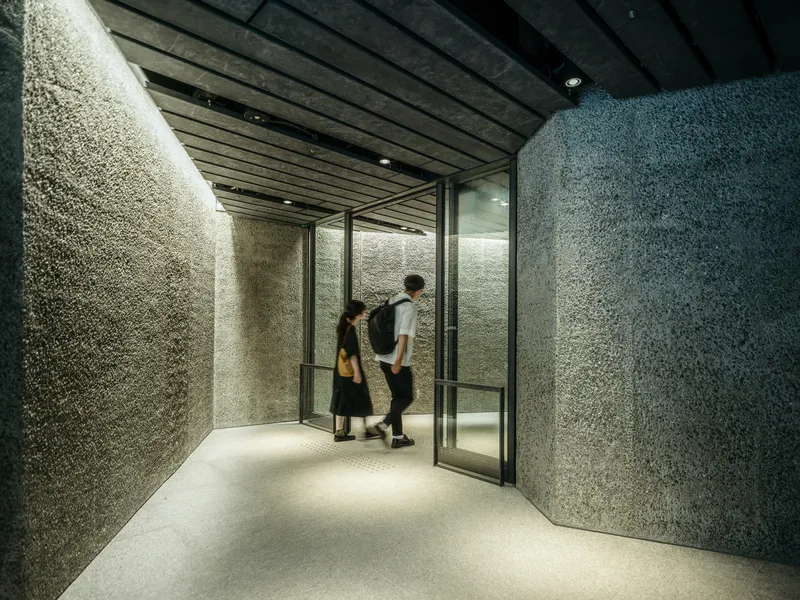 Corridor with textured aggregate walls and timber-clad ceiling as two visitors pass through the glazed doorway