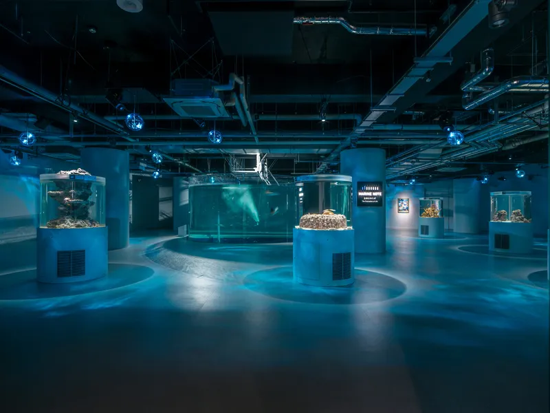 Interior exhibition space with cylindrical aquarium tanks under exposed mechanical ducts and blue lighting