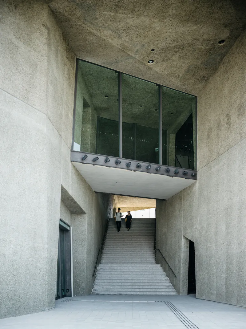 Concrete staircase ascending under a suspended glass box with two visitors climbing the steps