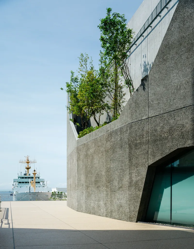 Concrete terrace with planted trees overlooking a harbor with a naval vessel in the distance