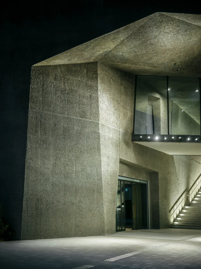 Textured concrete entrance canopy with glass walls and recessed lighting at night
