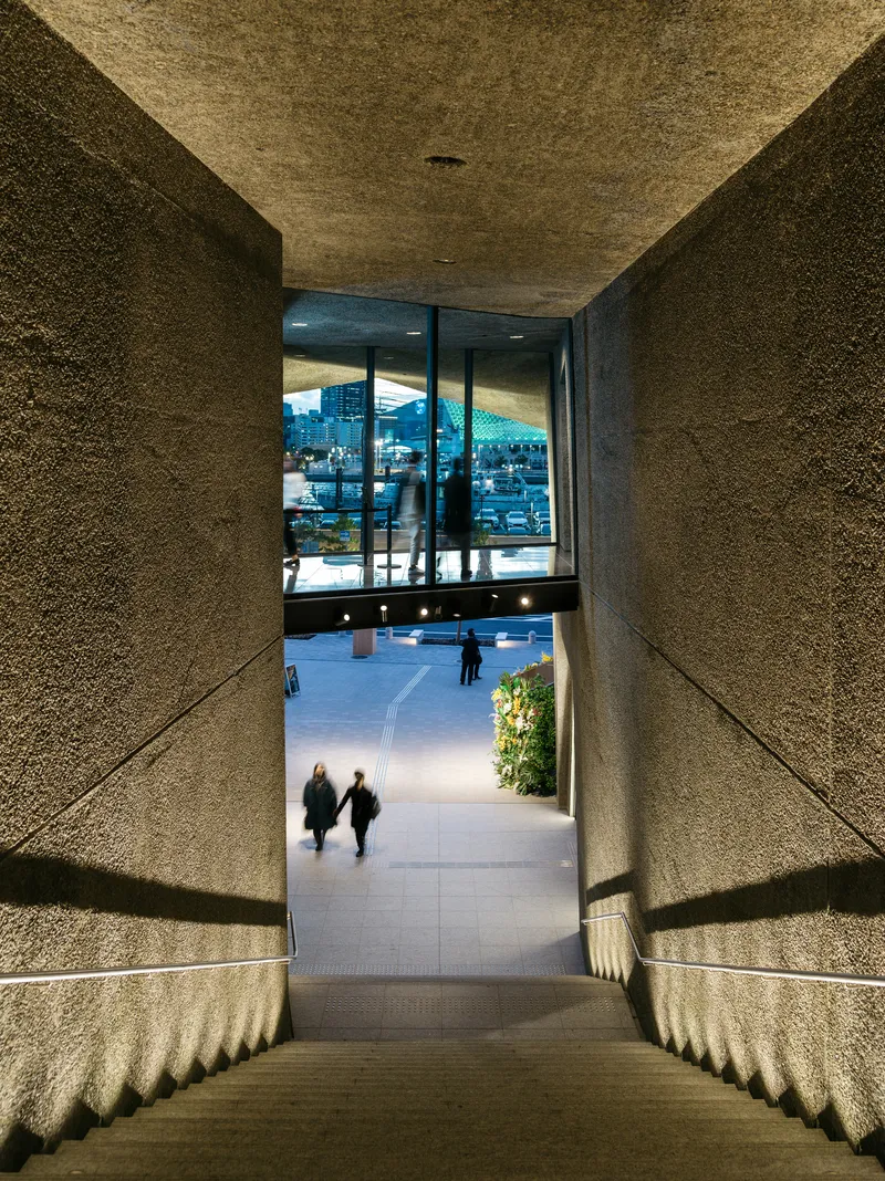View down the stepped concrete passage with textured walls framing visitors walking toward the plaza at dusk