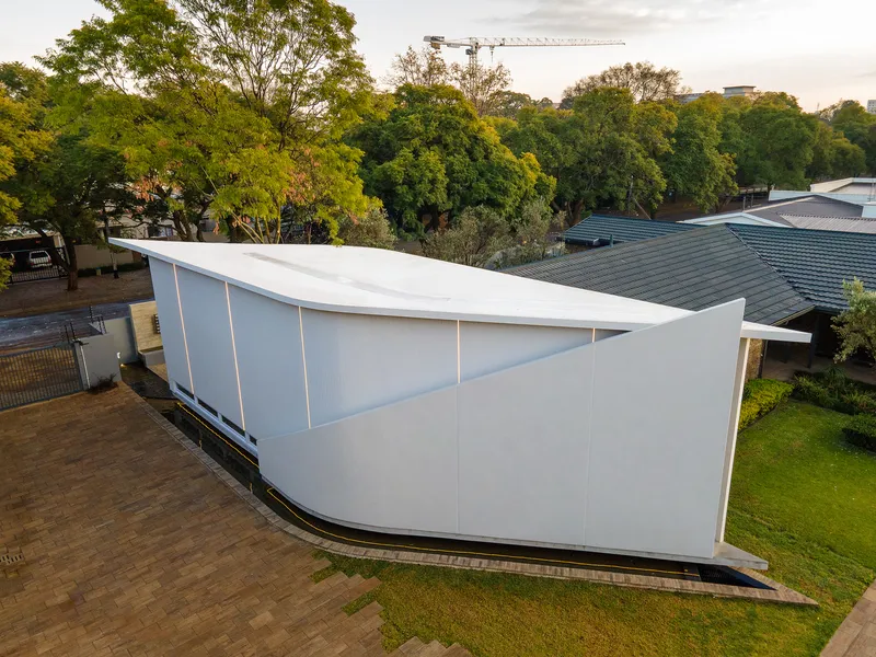Aerial view of the white curved volume with sloping roof set among mature trees at dusk