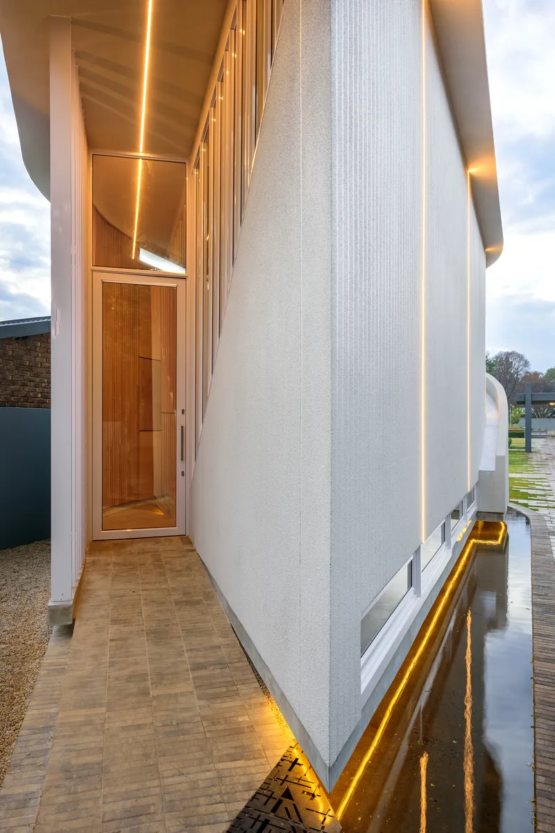 Covered entry passage with vertical ribbed cladding, timber door and linear lighting reflecting in wet pavement