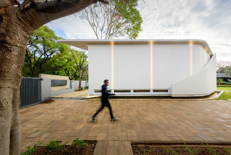 White ribbed facade with vertical light slots as a pedestrian crosses the brick forecourt beneath large trees
