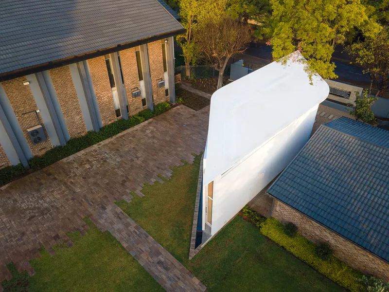 Curved white facade volume standing between brick paver courtyard and neighboring tiled roof buildings at dusk