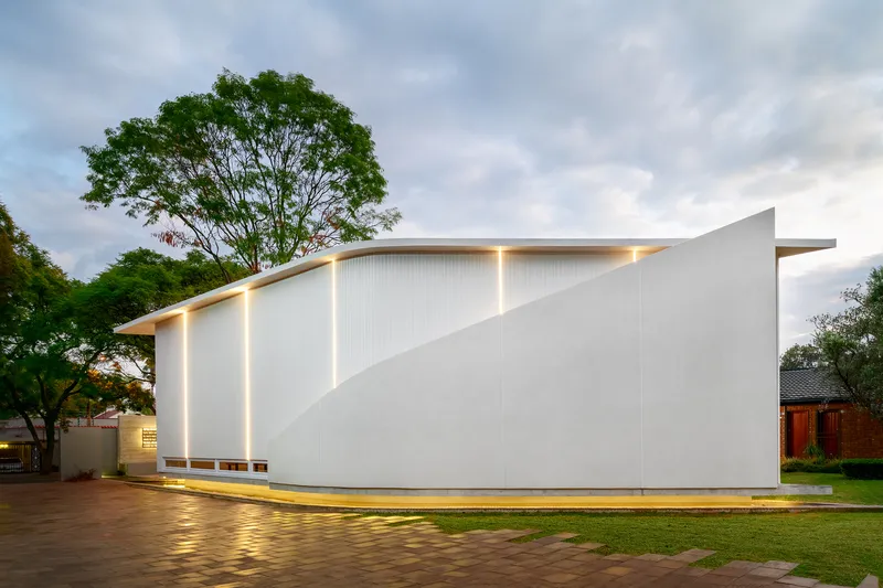 White corrugated metal facade with angled roofline and vertical light slots at twilight beside mature trees